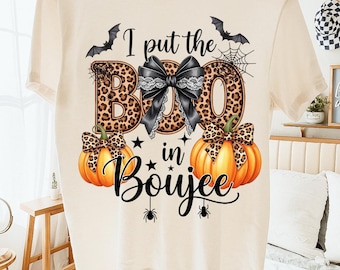 Może przedstawiać: Kremowa koszulka z motywem Halloween. Projekt zawiera słowa "I put the BOO in Boujee" z literami w panterkę, czarną kokardę, dynie, nietoperze, pająki i gwiazdy. Koszulka wisi na drewnianym wieszaku.