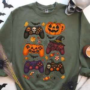 May include: Green sweatshirt featuring a Halloween-themed design. The design includes several game controllers with Halloween elements like pumpkins, bats, and a witch's hat. Candy and autumn leaves are also included in the design.