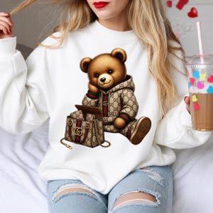 May include: A white sweatshirt featuring a brown teddy bear wearing a designer hoodie and matching shoes. The bear is seated next to a designer handbag. The bear and handbag have a repeating logo pattern in brown, green, and red.