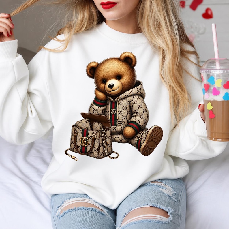 May include: A white sweatshirt featuring a brown teddy bear wearing a designer hoodie and matching shoes. The bear is seated next to a designer handbag. The bear and handbag have a repeating logo pattern in brown, green, and red.