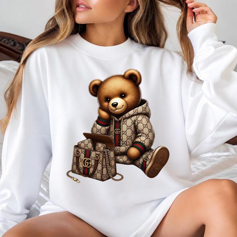May include: White sweatshirt featuring a brown teddy bear wearing a patterned hoodie and matching shoes. The bear is seated next to a matching handbag with a gold chain strap. The design incorporates brown, green, and red accents.