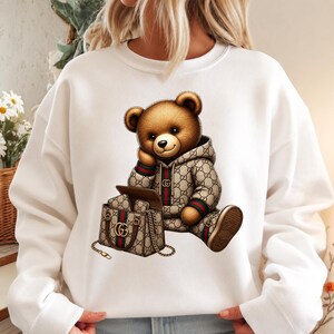 May include: White sweatshirt featuring a brown teddy bear wearing a patterned hoodie and shoes. The bear is sitting next to a matching handbag with a gold chain. The design incorporates green and red stripes.