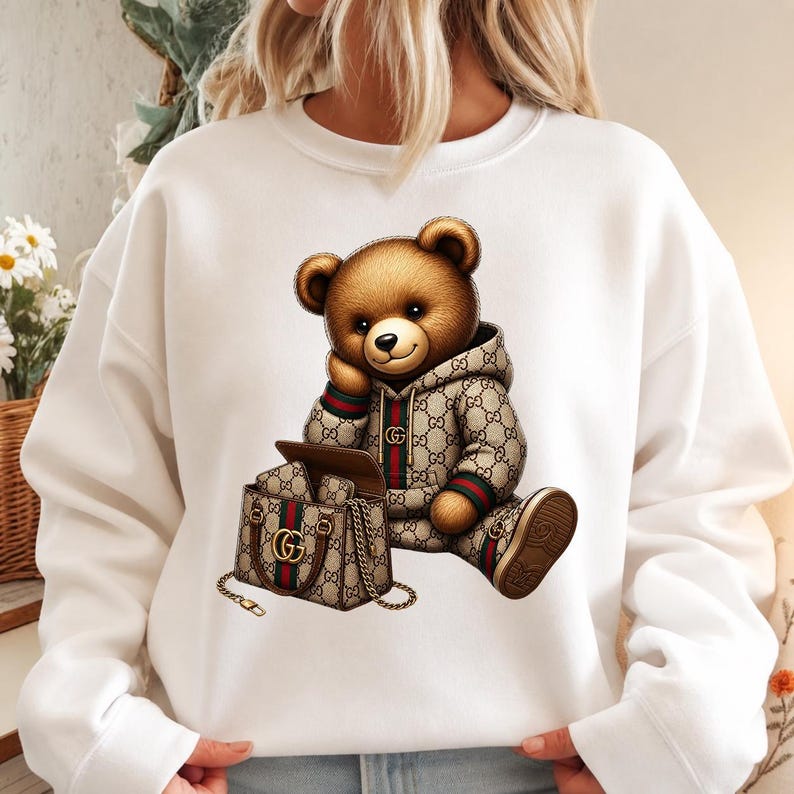 May include: White sweatshirt featuring a brown teddy bear wearing a patterned hoodie and shoes. The bear is sitting next to a matching handbag with a gold chain. The design incorporates green and red stripes.