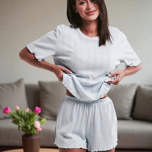 Pode incluir: Conjunto de pijama de malha canelada azul claro. O conjunto inclui uma blusa de manga curta e cal&ccedil;&otilde;es a condizer com uma borda ondulada. A modelo est&aacute; a usar o conjunto num ambiente dom&eacute;stico. Um vaso de flores cor-de-rosa est&aacute; numa mesa de madeira.