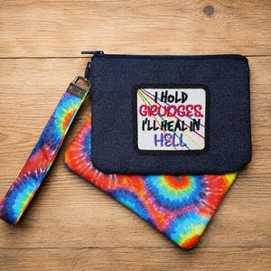 Op de afbeelding: Een donkerblauw denim tasje met rits en een polsband met een levendig regenboog tie-dye patroon. Een wit rechthoekig embleem is in het midden van het tasje geplaatst met de geborduurde tekst I HOLD GRUDGES. I'LL HEAL IN HELL in zwart, rood en paars.