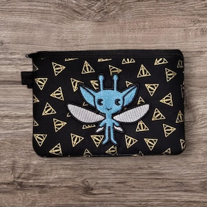 May include: Black zippered pouch with a repeating gold triangle pattern. A blue cartoon creature with wings is embroidered on the front. The pouch has a small black loop on the side.