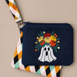 May include: A dark blue denim zippered pouch featuring an embroidered white ghost wearing a crown of red, orange, and yellow flowers. The pouch includes a wrist strap with a diamond pattern in orange, black, pink, and white, attached by a metal ring.