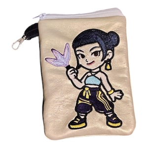 May include: A beige zippered pouch with a cartoon embroidered design. The design features a character with dark hair, a blue top, and dark pants with yellow accents. The character is holding a purple object. The pouch has a silver zipper and a black wrist strap.