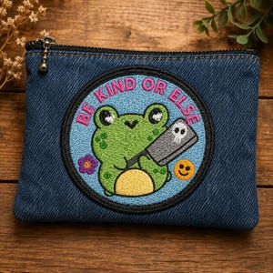 Op de afbeelding: Een donkerblauwe denim etui met rits en een ronde geborduurde patch. De patch toont een groene kikker die een grijs hakmes met een schedelpictogram vasthoudt, een kleine paarse bloem, een geel smileygezicht en de tekst BE KIND OR ELSE.