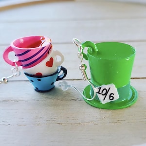 May include: Whimsical earrings featuring a stack of three miniature teacups in pink, white, and blue, and a green top hat with a small tag that reads "10/6". The earrings are designed with silver-colored hooks.