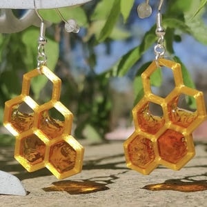 Pode incluir: Um par de brincos dourados em forma de favo de mel com ganchos prateados. Cada brinco apresenta um design de favo de mel tridimensional com resina âmbar preenchendo as células hexagonais. Os brincos estão suspensos em um fundo desfocado de folhagem verde.