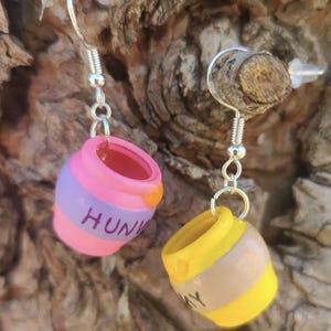 May include: A pair of whimsical earrings featuring miniature honey pots. One pot is pink and purple with "HUNNY" written on it, the other is yellow and tan with "HUNNY" written on it. The earrings have silver-colored hooks.