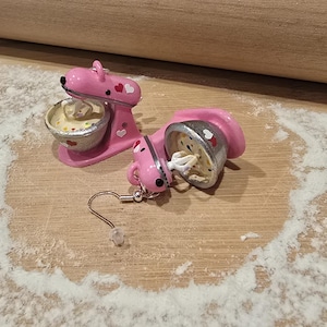 Può includere: Orecchini a forma di impastatrice rosa con dettagli a cuore, uno sdraiato su un lato. Gli orecchini sono su una superficie di legno cosparsa di farina, vicino a un mattarello.