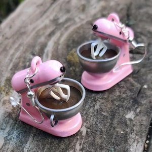May include: A pair of earrings shaped like miniature pink stand mixers. Each mixer features a silver-toned bowl filled with a dark brown substance and a white whisk attachment. The earrings include silver-toned metal hooks for attachment.