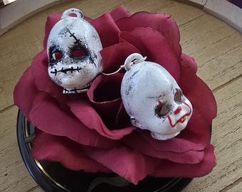 May include: A pair of earrings featuring two small, white doll heads with painted facial details. One head has black stitched markings over the eyes and mouth, while the other features red painted lips and dark eye makeup. Both heads have silver-toned hooks.