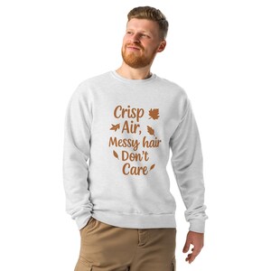 May include: A light gray sweatshirt with the text "Crisp Air, Messy hair Don't Care" in brown. The design includes leaf illustrations. The person in the image is wearing khaki pants.