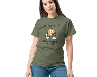Puede incluir: Camiseta verde oliva con el texto "I attract success, mostly by accident, but still." La camiseta presenta un personaje de dibujos animados sosteniendo dinero. La modelo lleva vaqueros azules.