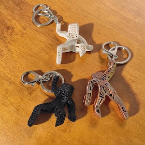 Puede incluir: Tres llaveros con herrajes plateados y colgantes abstractos con forma de figura humana. Uno es blanco, otro negro y el otro color cobre. Los colgantes tienen un diseño recortado. Los llaveros están sobre una superficie de madera.