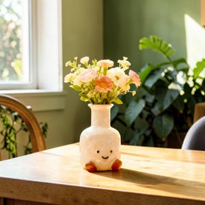 Puede incluir: Un jarrón de cerámica blanco con una cara sonriente y pequeños pies naranjas, que contiene un ramo de flores rosas, naranjas y blancas. El jarrón está sobre una mesa de madera, con una ventana y vegetación en el fondo.