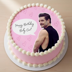 May include: A round pink cake with white icing and a decorative border. The cake features an image of a man and the text "Happy Birthday Baby Girl" in a speech bubble, with small heart shapes scattered across the top.