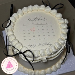 Puede incluir: Una tarta blanca de dos pisos con glaseado de crema y lazos negros. La capa superior presenta un diseño de calendario para octubre, resaltando el día 8 con un corazón. Debajo están las palabras "Happy Birthday".