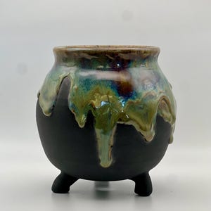 May include: A small, round ceramic cauldron with three short legs. The base is matte black, while the upper portion features a glossy, dripping glaze in shades of teal, green, and brown that cascades down the sides toward the center of the vessel.