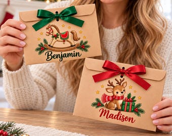 Puede incluir: Dos sobres rectangulares de papel kraft con ilustraciones navideñas. Un sobre muestra un caballito de madera con un lazo verde y el nombre Benjamin. El otro muestra un reno con un lazo rojo y el nombre Madison.