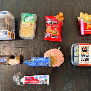 May include: A collection of novelty food items, including a Funko Pop figure, a bag of "Gory-Toes" chips, and a container of "Spewi Sushi". Other items include a candy bar, a tube of toothpaste, and a container of french fries.
