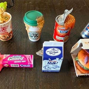 May include: A collection of novelty food items, including a cup of noodles, a coffee cup, a can of soup, a soda can, a pink blob with gum, a carton of milk, and a burger in a box. The burger box has the text "The Bugs are Better at Bug King!"