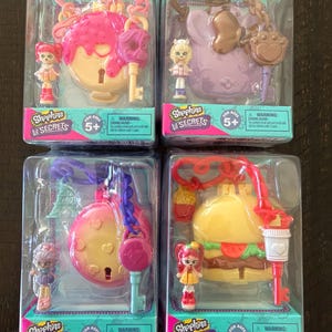 May include: Four Shopkins Li'l Secrets toy sets in clear plastic packaging. Each set includes a small figurine and a themed lock with a key. The locks are shaped like a donut, a bunny, a heart, and a burger. The packaging includes the text "Shopkins Li'l Secrets" and "For Ages 5+".