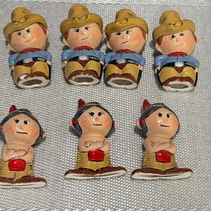 May include: A collection of seven small ceramic figurines arranged in two rows. The top row features four figures wearing tan cowboy hats, blue shirts, and tan pants. The bottom row features three figures with dark hair, red accents, and tan clothing.