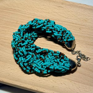 May include: A braided bracelet crafted from numerous small turquoise, brown, and black seed beads. The bracelet features silver-toned metal end caps and a silver-toned lobster claw clasp with an adjustable chain extension for a secure fit.