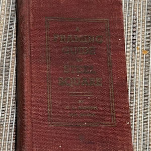 May include: A vintage, maroon-colored book titled "A Framing Guide to the Steel Square" by W.L. Higdon. The book has gold lettering and is slightly worn, suggesting age. The book is resting on a textured, neutral-toned surface.