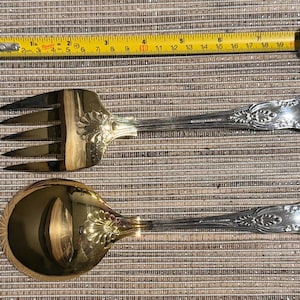 May include: A silver-toned salad fork and spoon set with ornate floral detailing on the handles. The fork has three tines, and the spoon has a rounded bowl. A yellow measuring tape is in the background.