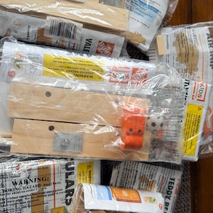May include: Several sealed packages containing wooden craft kits. The kits include wooden pieces, orange plastic components, and printed instructions. Some packages have warning labels about choking hazards and small parts.