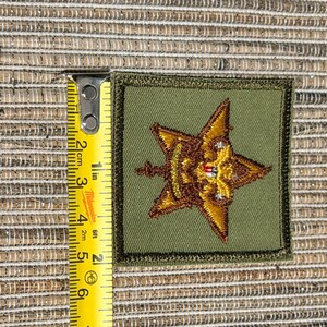 May include: A square olive green embroidered patch featuring a gold star emblem with a small red and white detail in the center. A yellow Milwaukee measuring tape is placed along the left edge, showing the patch is approximately 2 inches tall.