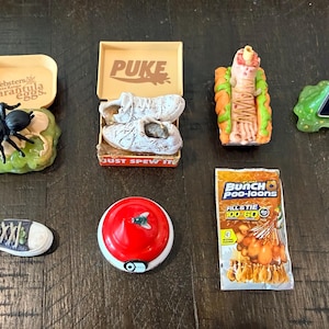 May include: A collection of novelty items, including a spider in a container labeled "Websters tarantula eggs", a pair of shoes in a box labeled "PUKE", a hot dog, a phone with "Sik Tok" on the screen, a shoe, a tomato, and a package of "Bunch poo-loons".