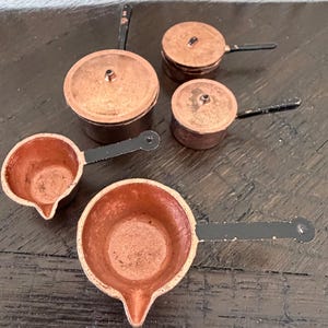 May include: A collection of miniature copper pots and pans. The set includes two saucepans with long black handles, two lidded pots, and a small pot with a lid. The copper cookware has a rustic, aged appearance, suggesting vintage or antique items.