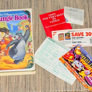 May include: A vintage VHS tape of Disney's "The Jungle Book" with colorful cover art featuring Baloo and Mowgli. Also includes promotional materials, a coupon for Teddy Grahams, and a flyer for "The Brave Little Toaster" video.