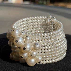 May include: A multi-strand cuff bracelet composed of numerous small, cream-colored faux pearls arranged in a wide, cylindrical band. One side of the bracelet features a cluster of larger, varying-sized cream-colored faux pearls with dark centers.