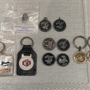 May include: A collection of snowmobile-themed accessories including a clear plastic Polaris keychain, a black leather Arctic Cat keychain, and several metal circular charms and keychains featuring embossed cat head logos in various metallic finishes.
