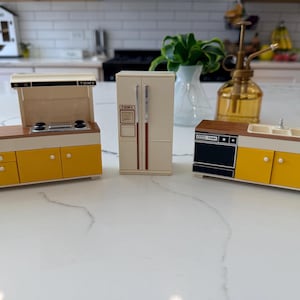 May include: A set of vintage toy kitchen furniture, including a stove, refrigerator, and sink unit. The pieces are primarily yellow and cream with brown accents. The stove has a range hood, and the refrigerator has a TOMY logo. The sink unit has a small faucet.