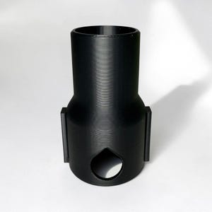 Może przedstawiać: Czarny adapter drukowany w 3D z cylindryczną g&oacute;rą i rozszerzoną podstawą. Adapter posiada otw&oacute;r w kształcie dziurki od klucza i dwa prostokątne boczne przedłużenia. Obiekt jest przeznaczony do konkretnego celu, prawdopodobnie do łączenia lub adaptacji komponent&oacute;w.