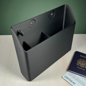 May include: A matte black organizer with two internal dividers and mounting holes. The organizer is rectangular with a slanted top and a small hook. A passport and a card are visible in the background.