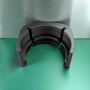 Puede incluir: Un componente de plástico negro semicircular con una textura acanalada. El objeto tiene un diseño curvo con múltiples capas apiladas, sobre un fondo verde y plateado.