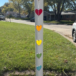 May include: A decorative outdoor hanging ornament featuring seven heart-shaped glass pieces in a rainbow of colors: red, orange, yellow, green, blue, and purple. The hearts are connected by a silver chain and hang from a white pole.