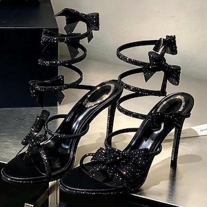 Könnte beinhalten: Ein Paar schwarze High-Heel-Sandalen mit spiralförmigem Knöchelriemen-Design, das mit funkelnden Kristallen verziert ist. Jeder Schuh verfügt über eine dekorative Schleife an der Zehenpartie und am Knöchelriemen sowie einen dünnen Stiletto-Absatz.