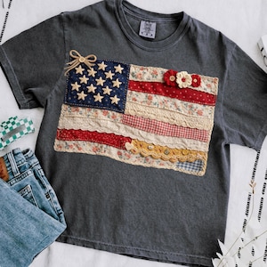 Puede incluir: Camiseta gris oscuro con un diseño de bandera americana de patchwork. La bandera presenta un campo azul con estrellas bordadas, rayas rojas y blancas y detalles florales y de encaje. Un pequeño lazo y adornos florales.