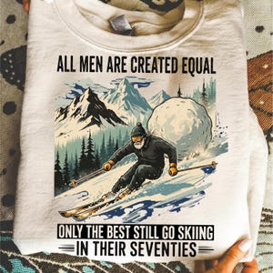 May include: Cream-colored sweatshirt with a graphic of a skier on a snowy mountain slope. The image includes text that reads "ALL MEN ARE CREATED EQUAL" and "ONLY THE BEST STILL GO SKIING IN THEIR SEVENTIES."