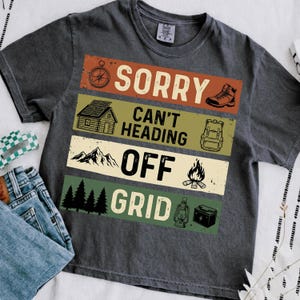 Op de afbeelding: Donkergrijs T-shirt met de tekst "SORRY CAN'T HEADING OFF GRID" in een gelaagd ontwerp. Het ontwerp bevat een kompas, wandelschoen, hut, rugzak, bergen, kampvuur, bomen, lantaarn en batterij.